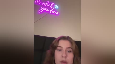 Media: A video of a young woman with long, wavy brown hair, wearing makeup and a black top, standing in a dimly lit room. Neon pink text on the wall reads, \"do what you love.\