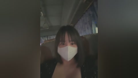 Media: A dimly lit video of an Asian woman with straight, shoulder-length black hair, wearing a white medical mask and a black blouse, seated indoors.