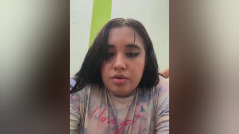 Media: Video of a young Latina girl with long, dark hair, wearing a pastel-colored t-shirt with \"I love you\" written in red cursive. She has a neutral expression, and the background features a lime-green wall with polka dots.