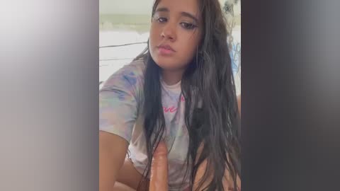 Media: Video of a young woman with long dark hair, wearing a tie-dye shirt, looking down. Her expression is neutral.