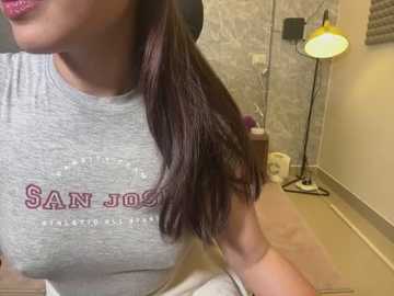 Media: Video of a woman with long, dark hair in a ponytail, wearing a gray San Jose State University t-shirt, seated indoors with beige walls, a lamp, and a towel rack in the background.