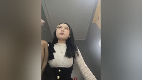 Media: Video of an Asian woman with long black hair, fair skin, wearing a white sweater and black vest, looking up, in a dimly lit corridor.