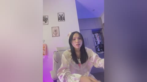 Media: Video of an Asian woman with long black hair, wearing a white robe, sitting on a white chair in a dimly lit room with purple lighting. Framed black-and-white art pieces hang on the wall.