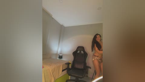 Media: A video of a small, sparsely furnished bedroom with beige walls, a single bed with a beige sheet, a black gaming chair, and a smiling young woman in lingerie.