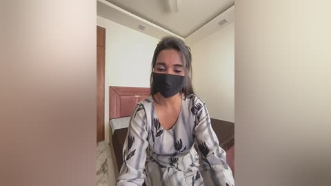 Media: Video of a woman with medium brown skin, wearing a black face mask, white floral-patterned robe, and sitting in a beige room with a wooden bed and white walls.