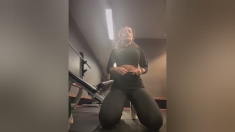 Media: Video of a fit Caucasian woman with long blonde hair kneeling in a dimly lit gym. She wears a black sports bra and leggings, looking confident. Equipment and workout benches in the background.