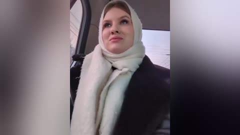 Media: A video of a woman with fair skin, wearing a white hijab and a dark coat, seated in a car with the driver's side window visible.