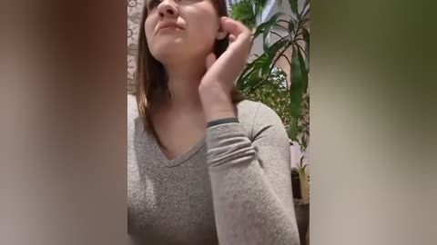 Media: Video of a woman with light skin and brown hair, wearing a gray sweater, leaning against a green plant, in a home setting.