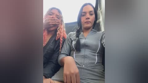 Media: Video of two women, one with pink dreadlocks and a black jacket, the other with long black hair in a braid, wearing a grey zip-up hoodie, sitting in a car.