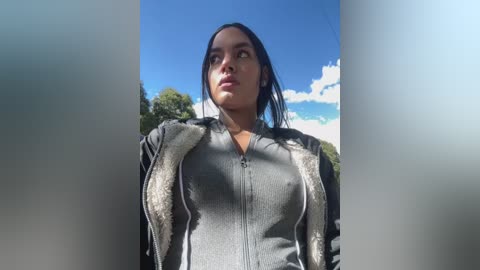 Media: A video of a Latina woman with long black hair, wearing a grey zip-up hoodie, standing outdoors under a bright blue sky with white clouds and green trees in the background.