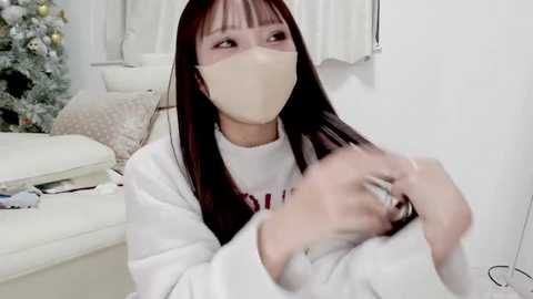 Media: Video of an Asian woman with long black hair, wearing a white mask, a white sweater, and gloves, sitting on a white couch in a minimalist living room with a Christmas tree in the background.