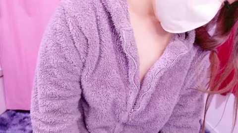 Media: Video of a person wearing a lavender, plush bathrobe and a white face mask, with a pink towel and purple carpet in the background.