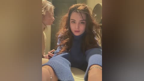 Media: Video of a young woman with curly brown hair, wearing a blue turtleneck dress, seated in a dimly lit bathroom, looking at an elderly man with white hair.