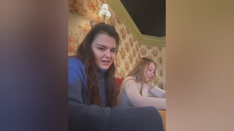 Media: Video of two young women, one with long dark hair in a blue sweater, the other with light brown hair in a white top, sitting on a couch in a floral wallpapered room.