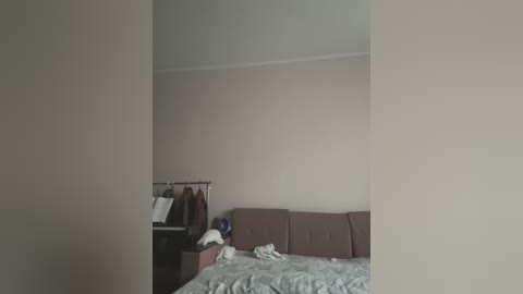 Media: A video of a minimalist bedroom with a beige wall, a brown couch, and a cluttered dark wooden nightstand with a hanger and clothes. The bed has a white and gray patterned duvet.