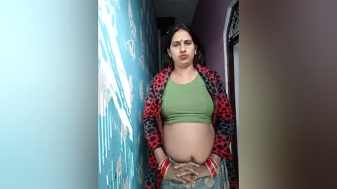 Media: Video of a pregnant South Asian woman with medium-brown skin, wearing a green crop top, red polka-dot jacket, and silver bangles, standing in a narrow hallway with blue and purple walls.