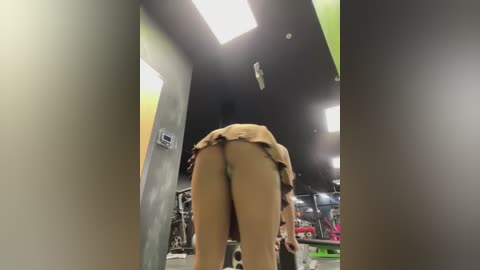 Media: Video of a woman bending over, exposing her bare buttocks in a gym setting. She wears a brown skirt lifted above her waist, revealing her nude lower body. Background includes gym equipment and green wall.