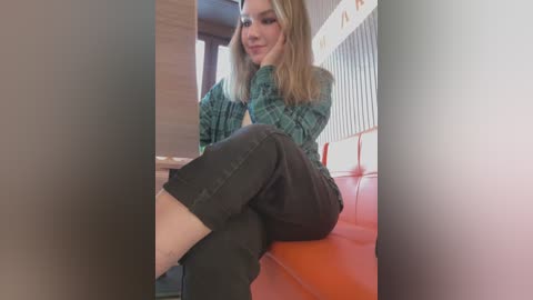 Media: Video of a blonde woman with fair skin and medium build, wearing a green plaid sweater and dark jeans, sitting on a red bench in a modern room with wooden and metal accents.