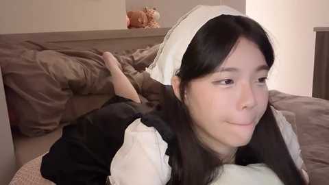 Media: Video of an Asian woman lying on a bed in a maid outfit, black dress with white apron and headpiece, smiling, brown bedding, stuffed toy in background.