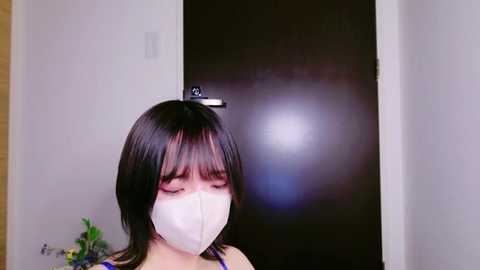 Media: Video of an Asian woman with straight black hair, wearing a white face mask, standing in a dimly lit hallway.