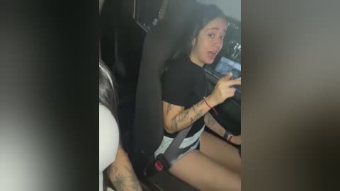 Media: Video of a tattooed woman wearing a black shirt and gray shorts, driving a car, with a blurred background.