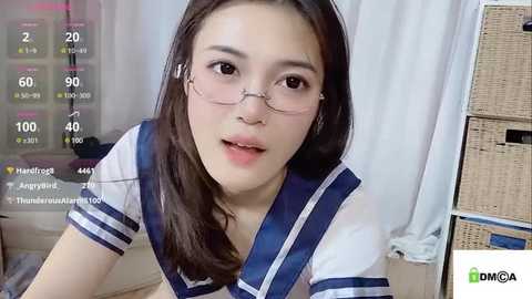 Media: A video of a young Asian woman with fair skin, long dark hair, and glasses, wearing a sailor-style school uniform, indoors with a white curtain background, recorded on a smartphone.