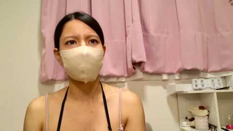 Media: Video of an East Asian woman with fair skin and dark hair, wearing a white face mask and a black spaghetti strap top. Background features light pink curtains and a white shelf with various items.