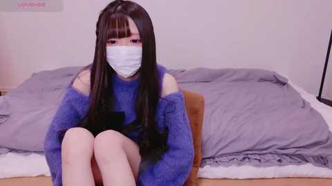 Media: A young Asian woman with long black hair, wearing a blue knit sweater and white surgical mask, sits on a bed with grey sheets and a white pillow in a simple, clean bedroom.