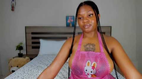 Media: Video of a dark-skinned woman with long braids, wearing a pink checkered apron with a cartoon bunny, sitting on a bed in a simple, beige room.