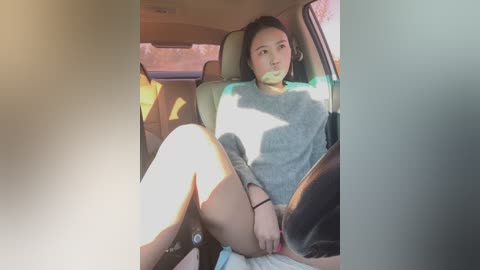 Media: Video of a young Asian woman with medium skin tone and dark hair, wearing a gray sweater, sitting in a car, with sunlight streaming through the windows, creating shadows.