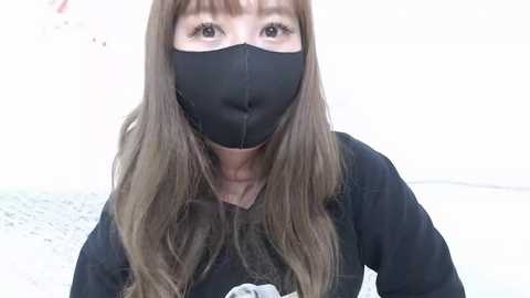 Media: Video of a person with long brown hair, wearing a black face mask and a black jacket, standing in front of a white wall, with a blurred background.