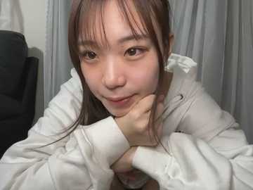 Media: Video of an East Asian woman with long brown hair, wearing a white hoodie, leaning forward with her chin on her hands, looking directly at the camera. Background features a gray curtain and a black leather chair.