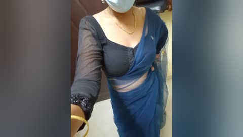 Media: Video of a woman with medium skin tone, wearing a black blouse and blue sari with a sheer black overlay, white face mask, and gold necklace, standing indoors.