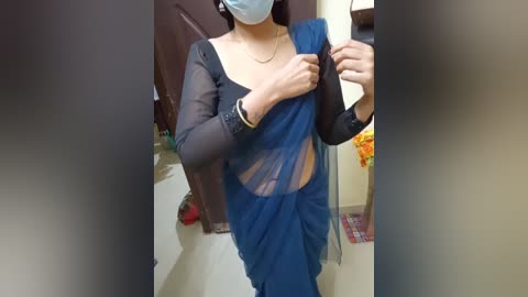 Media: Video of an Indian woman in a blue saree, black long-sleeve blouse, and blue face mask, adjusting her saree in a dimly-lit room with beige walls and a red chair.