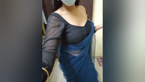 Media: Video of a woman with medium skin tone, wearing a black blouse and a blue saree, with a face mask, taken indoors.