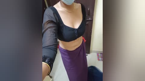 Media: Video of a light-skinned woman with a medium build, wearing a black, sheer, long-sleeved blouse and a purple wrap skirt. She has a blue face mask on, standing in a tiled room with a dark wooden door and colorful floor tiles in the background.