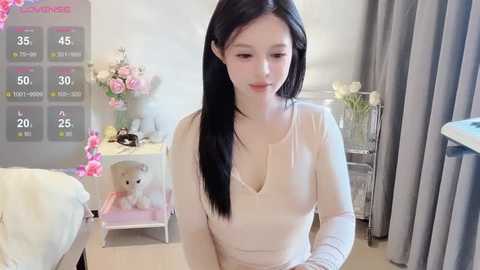 Media: Video of an East Asian woman with long black hair, light skin, and small breasts, wearing a beige long-sleeve top, kneeling on a carpeted floor. Background includes a bed, a flower vase, a weather display, and a cat.