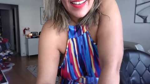 Media: Video of a smiling woman with shoulder-length blonde hair, wearing a colorful striped halter top, standing indoors with a modern, minimalist decor background.