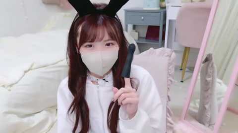 Media: A video of an Asian woman with long brown hair, wearing a white bunny costume with a face mask, holding a lollipop, in a cozy, pastel-colored room with a bed and pink furniture.