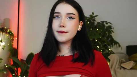 Media: Video of a young woman with pale skin, long black hair, and blue eyes, wearing a red top, standing in a room with string lights, green plants, and a couch.