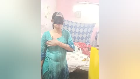 Media: Video of a woman in a teal, long-sleeved dress with a black head-mounted display, standing in a brightly lit bedroom with pink walls, a blue-patterned bedspread, and a white wall-mounted light.