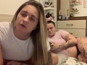 Media: Video of a pregnant woman with blonde hair, tattoos, and a white shirt, resting on a bed, while a woman with dark hair and a pink shirt looks at her phone in a cluttered bedroom.