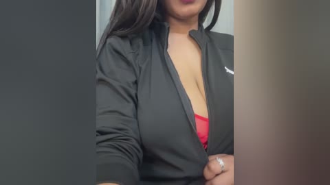 Media: Video of a woman with medium skin tone, wearing a black blazer open to reveal a red bra, standing against a blurred background.