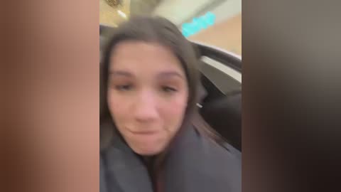 Media: A blurred video of a young woman with straight brown hair and fair skin, wearing a black jacket, sitting in a car. The background shows a blurred indoor setting with ambient lighting and blue lights.