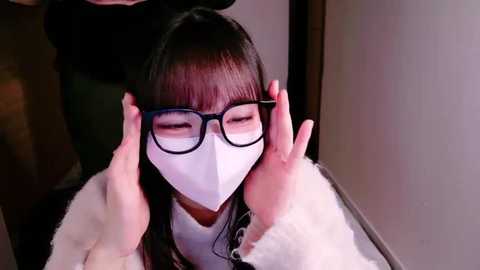 Media: Video of an Asian woman in a white mask and black-framed glasses, being held by hands, in a dimly-lit room with soft, warm lighting.