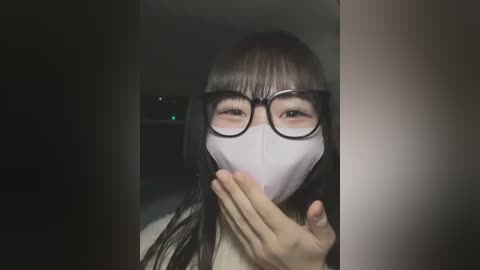 Media: Video of an East Asian woman with long black hair, wearing glasses and a white face mask, covering her mouth with her hand, set in a dimly lit room.