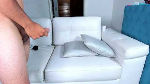 Media: Video of a nude man with a flaccid penis, standing beside a white leather sectional sofa with a gray pillow, in a modern, minimalist living room.