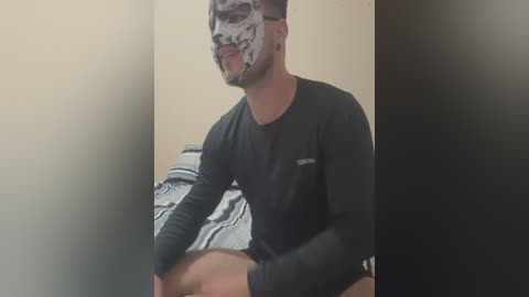 Media: Video of a young man with short dark hair wearing a black long-sleeve shirt, seated cross-legged on a bed with gray and white striped sheets. His face is partially covered with a white skull mask, creating a playful, Halloween-themed look.