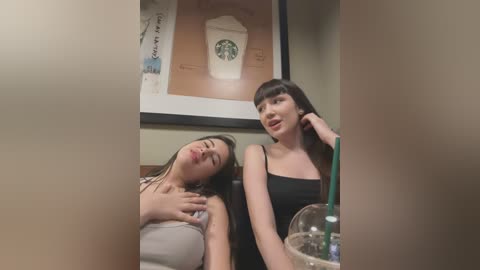 Media: Video of two young women with long hair, one with a tank top, one in a black top, seated at a table in a coffee shop, leaning on each other. Background includes framed Starbucks art and a green drink.