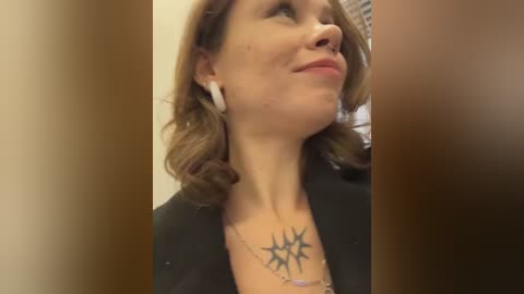 Media: A video of a fair-skinned woman with shoulder-length wavy brown hair, wearing a black jacket, and a star tattoo on her chest, looking upward with earbuds in.